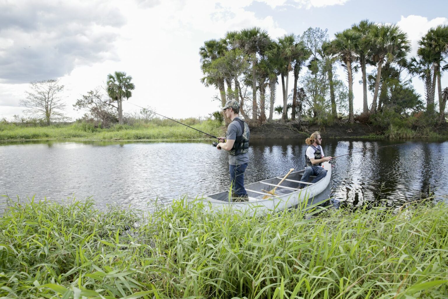 Royalty-free image - Two people fishing from a small boat on a calm river. They are surrounded by lush greenery and tall palm trees under a partly cloudy sky. Fishing from a canoe on the St. Johns River near Christmas, Florida.