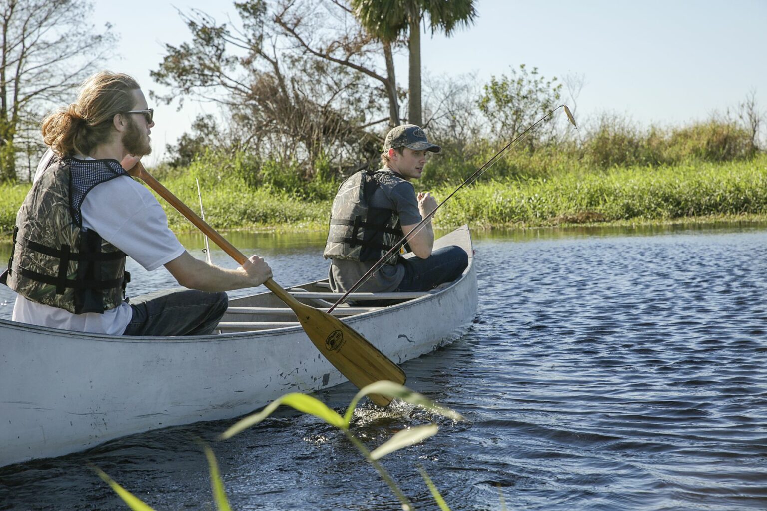 Royalty-free image - Two people wearing life vests paddle a canoe on a calm river. One holds a fishing rod while the other rows. The background includes green grass, palm trees, and clear blue skies. Fishing from a canoe on the St. Johns River near Christmas, Florida.