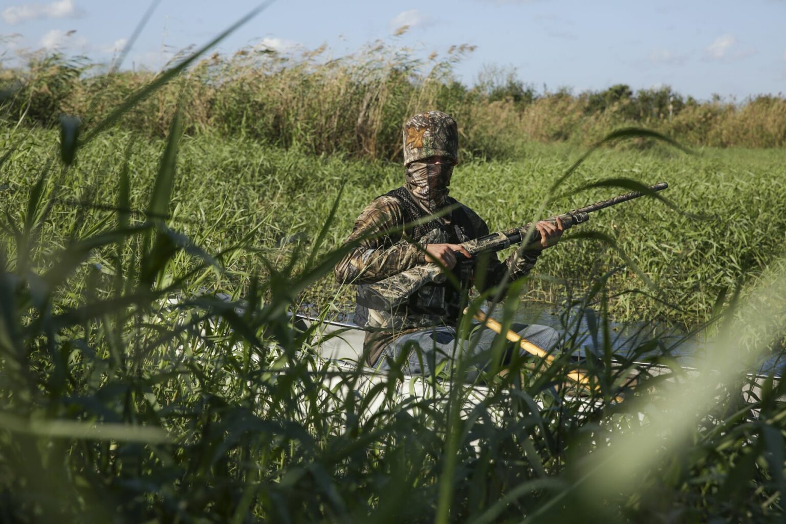 Royalty-free image - A person in camouflage gear is sitting in a kayak, holding a firearm amidst tall grass and reeds in a lush, natural setting. The sky is clear with a few clouds. Hunting from a canoe on the St. Johns River near Christmas, Florida.