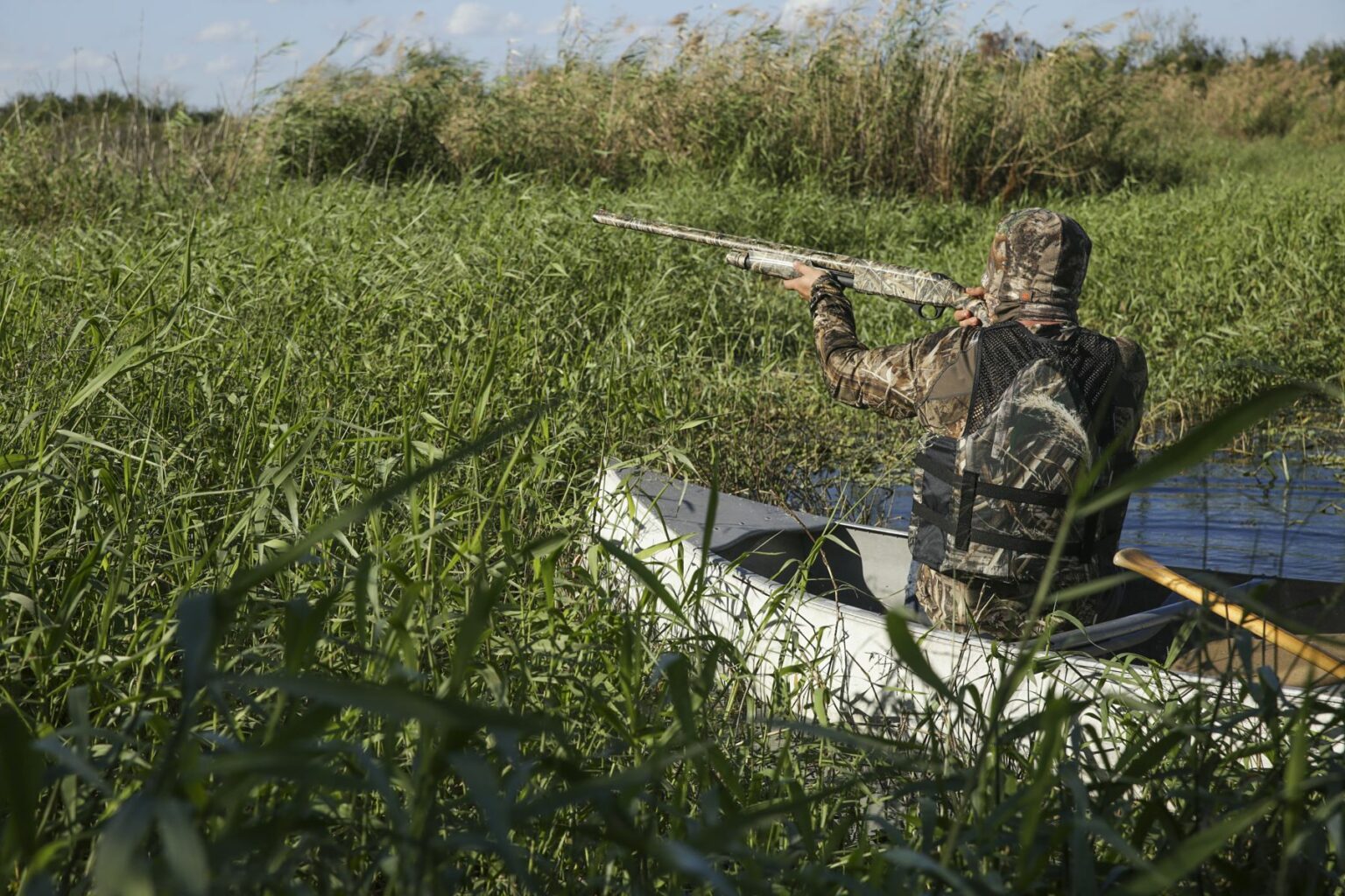 Royalty-free image - A person wearing camouflage clothing is sitting in a small boat, holding a shotgun and aiming into tall reeds and grass in a wetland area. The scene suggests hunting or birdwatching amidst a natural environment. Hunting from a canoe on the St. Johns River near Christmas, Florida.