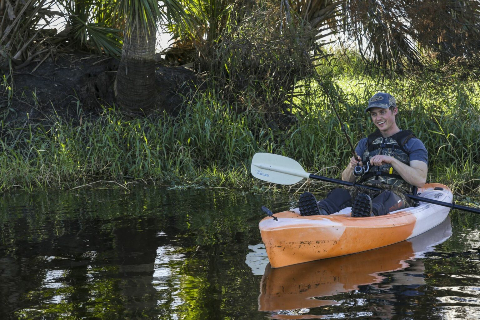 Royalty-free image - A man smiling while kayaking in a calm river, wearing a cap and life vest. He holds a fishing rod, surrounded by lush greenery and palm trees on the riverbank. The kayak is orange and the water reflects the scenery. Fishing from a kayak on the St. Johns River near Christmas, Florida.