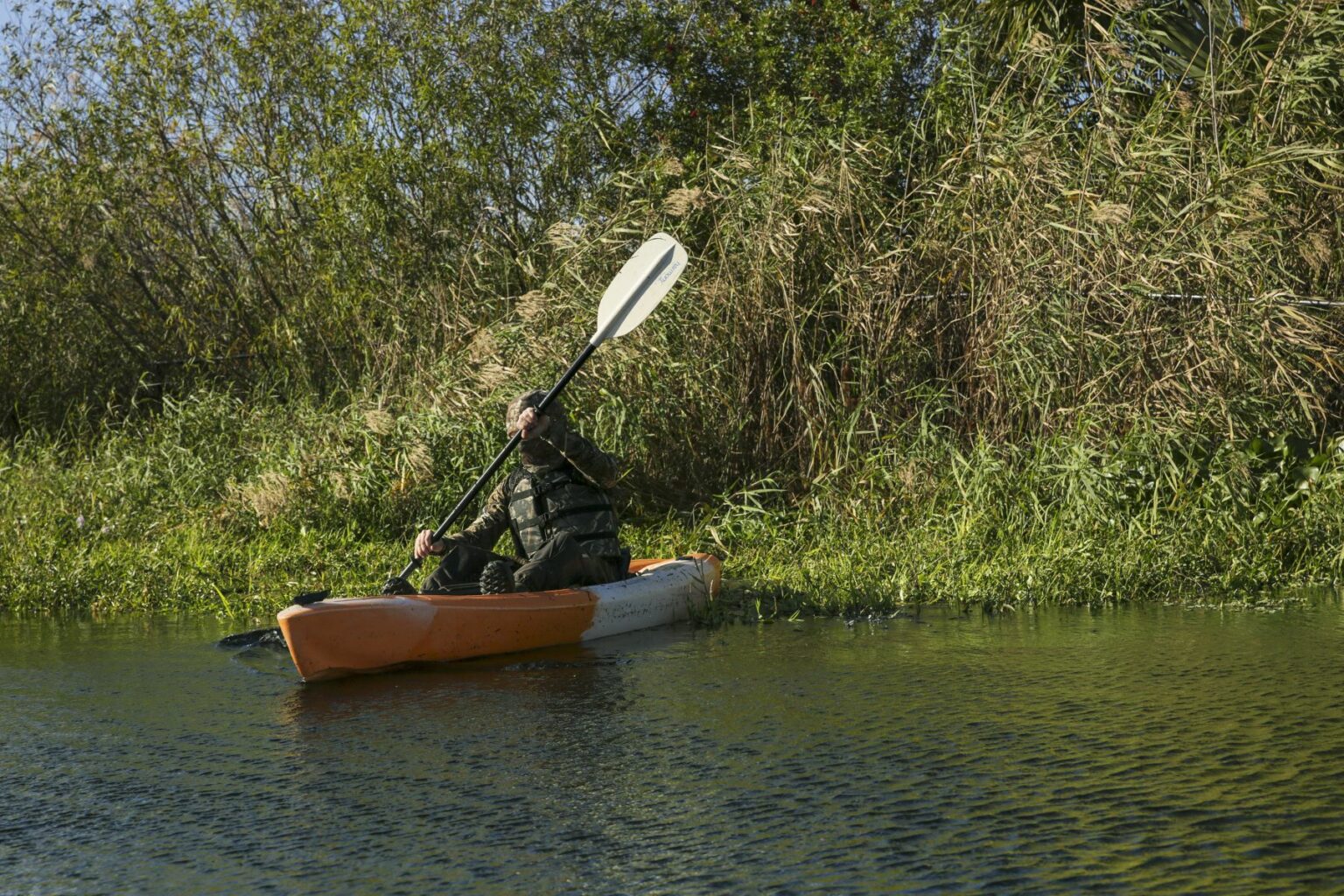 Royalty-free image - A person in camouflage attire is kayaking through a narrow waterway surrounded by lush greenery and tall reeds on a sunny day. The kayak is orange and white, and the individual is paddling along the calm water. Hunting from a kayak in the marsh along the St. Johns River near Christmas, Florida.