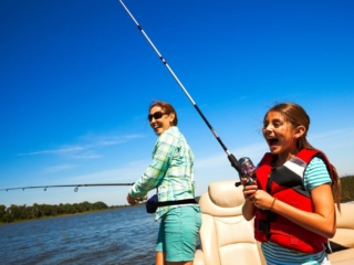 License-free image A woman and a young girl are fishing off the side of a boat. The woman is standing and casting a fishing rod, wearing sunglasses and a green shirt. The girl is wearing a red life jacket and patterned shorts, holding a fishing rod and smiling. Clear blue sky and trees in the background.
