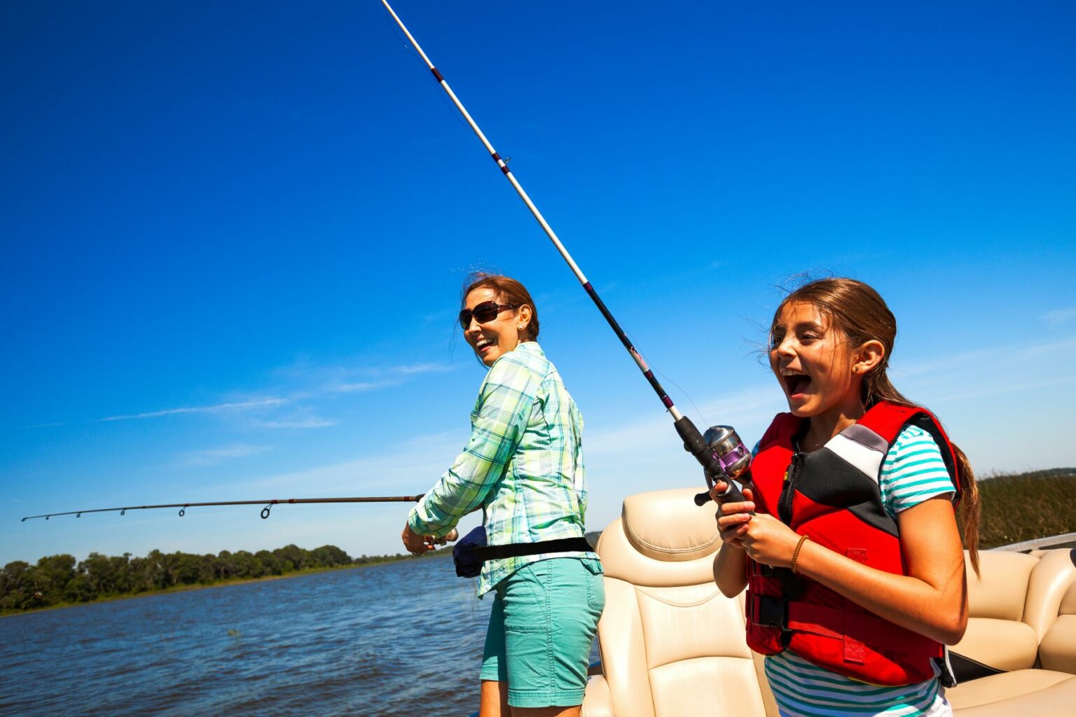 License-free image A woman and a young girl are fishing off the side of a boat. The woman is standing and casting a fishing rod, wearing sunglasses and a green shirt. The girl is wearing a red life jacket and patterned shorts, holding a fishing rod and smiling. Clear blue sky and trees in the background.