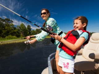 License-free image - A woman and a young girl are fishing off the side of a boat. The woman is standing and casting a fishing rod, wearing sunglasses and a green shirt. The girl is wearing a red life jacket and patterned shorts, holding a fishing rod and smiling. Clear blue sky and trees in the background.