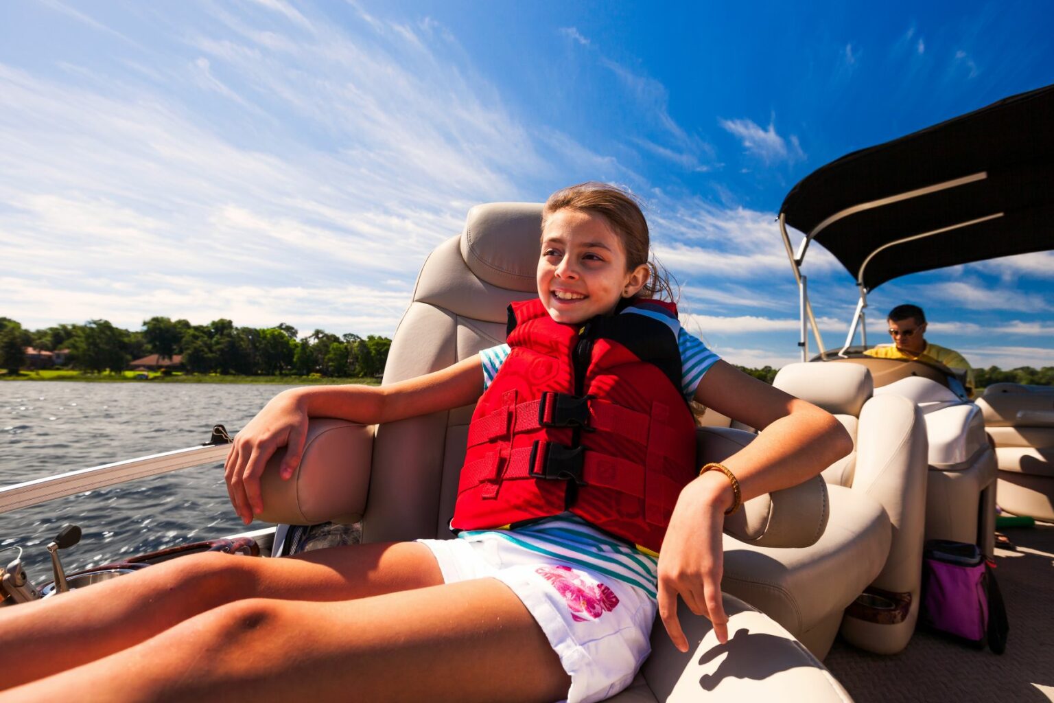 Royalty-free image - A girl wearing a red life vest and striped shirt is sitting comfortably on a boat, smiling. The sky is clear and blue, and there&#039;s water in the background. Another person is seated further back on the boat.