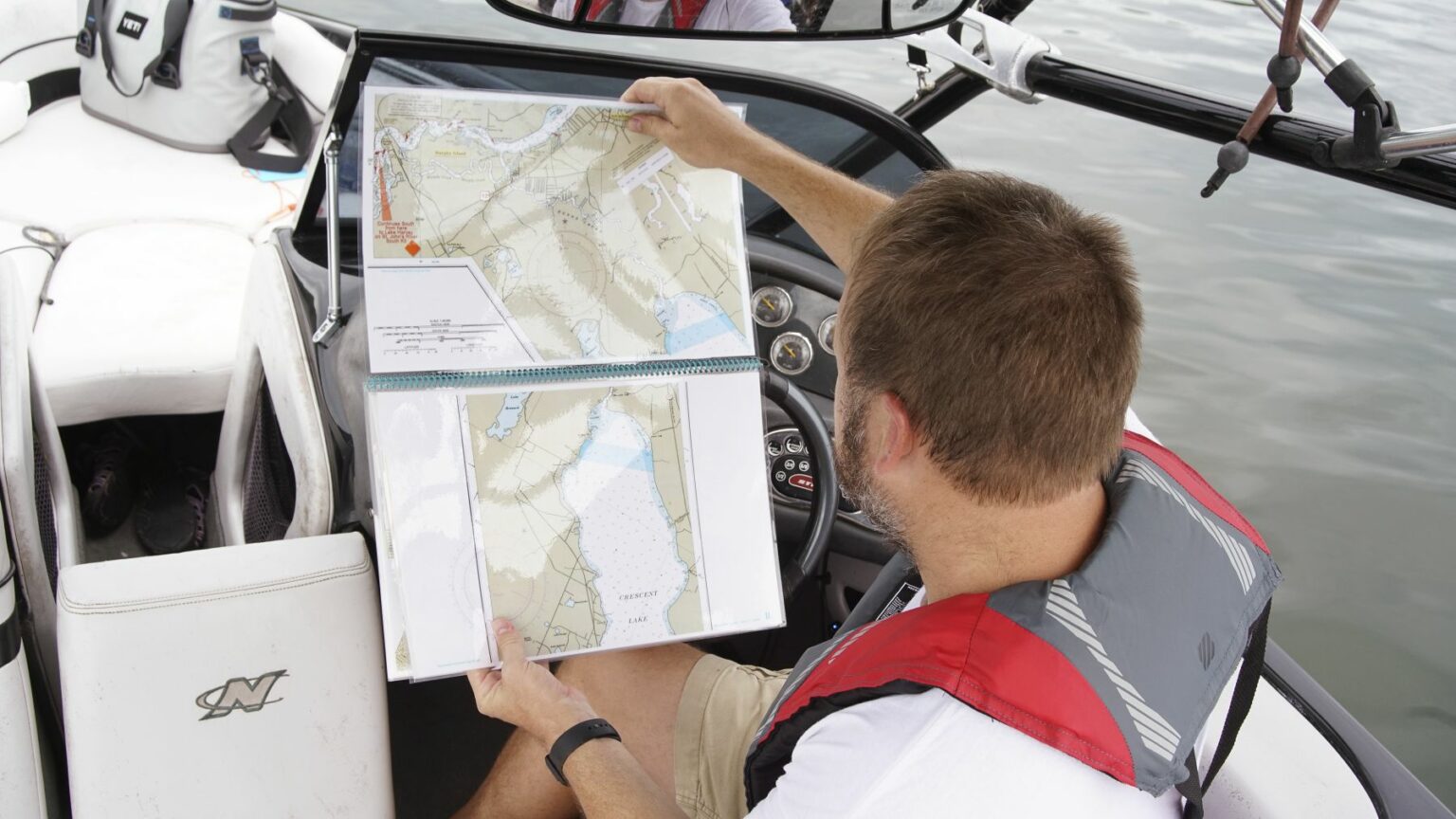 Royalty-free image - A man on a boat is looking at a map while wearing a life vest. He&#039;s sitting next to the steering wheel, and his focus is on the navigational charts in his hands. The boat is on still water.