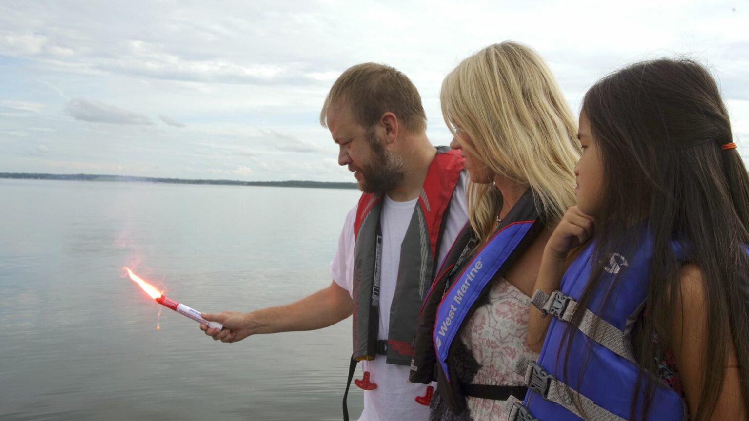 License-free image - A man holding a lit flare stands next to a blonde woman and a young girl with long dark hair. All wear life jackets and are on a boat with calm waters and a cloudy sky in the background. Using a flare during the day.