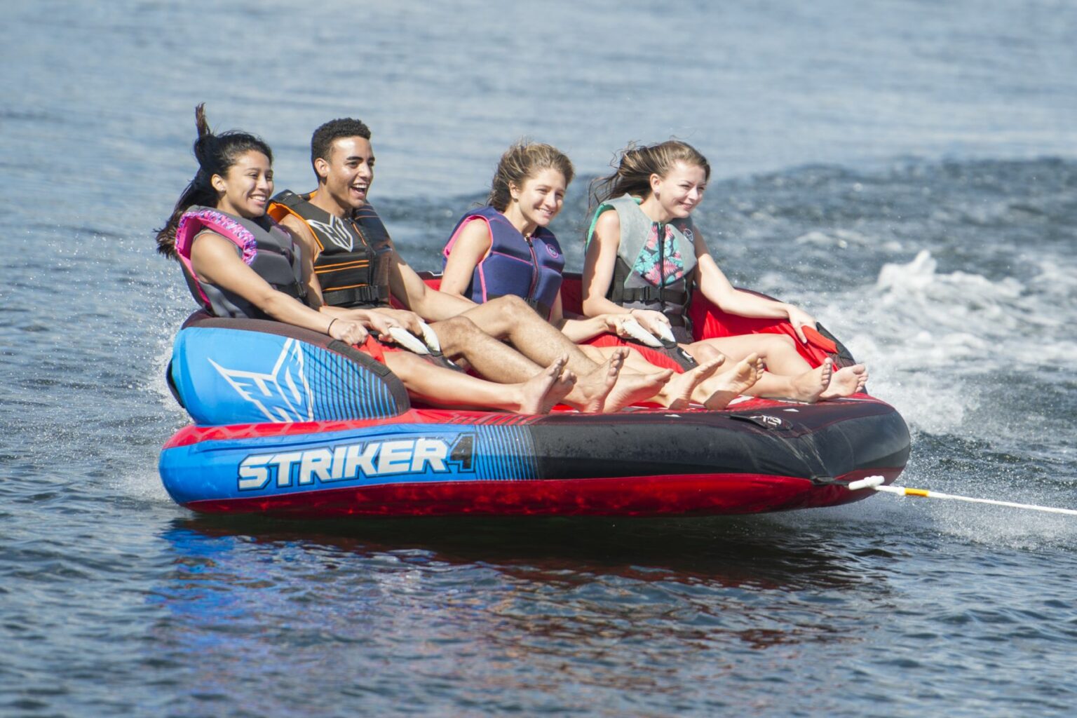 License-free image - Four people, wearing life vests, are sitting on an inflatable water tube labeled &quot;Striker 4.&quot; They are being towed across a body of water, creating a small wake behind them. Everyone appears to be enjoying the ride, smiling, and holding onto the tube&#039;s handles.