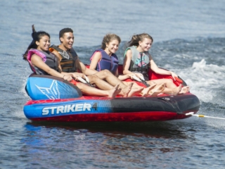 License-free image - Four people are enjoying a ride on an inflatable water tube labeled &quot;Striker 4.&quot; They are being pulled by a boat on a body of water, with smiles and laughter showing their excitement. All are wearing life jackets for safety.