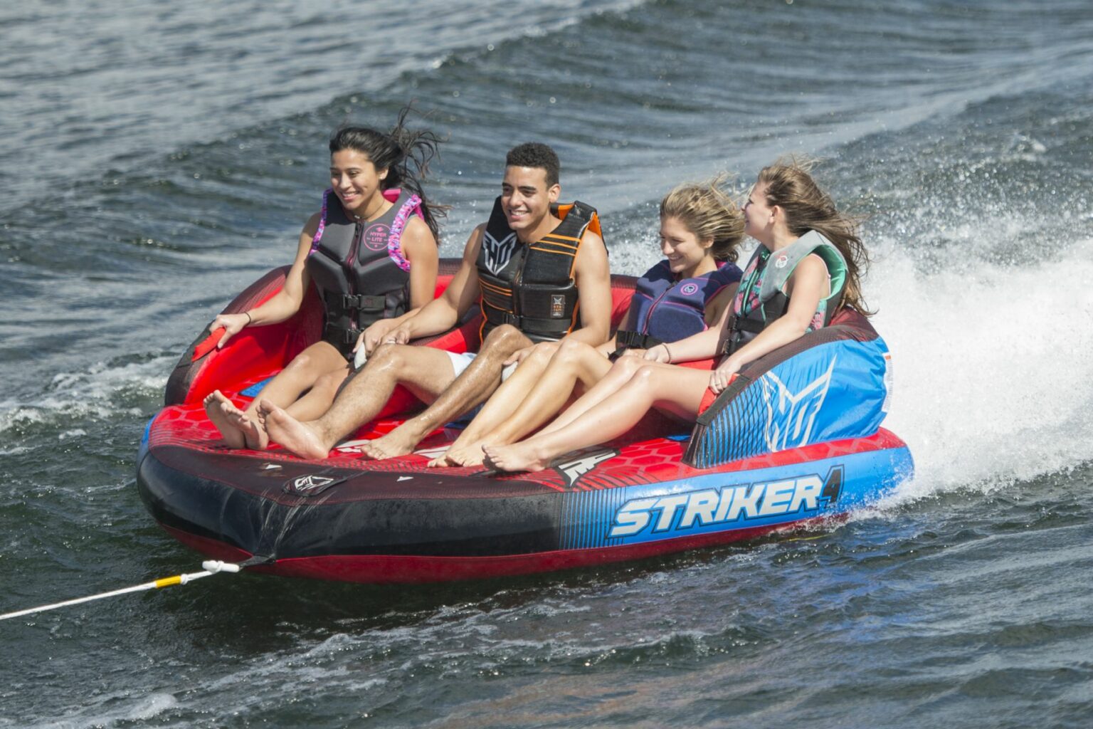 License-free image - Four people enjoy tubing on a large inflatable raft labeled &quot;Striker&quot; as it gets pulled across the water. They are smiling and holding on while wearing life jackets, with splashes and wake visible around the raft. The water is calm and blue under a sunny sky.