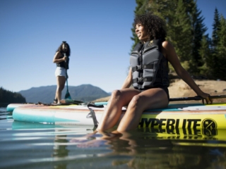 License-free image - Two women enjoy a sunny day on a lake. One is sitting on a paddleboard with her legs in the water, and the other is standing on a standup paddleboard (SUP) in the background. Both wear life vests, with mountains and trees visible in the background.