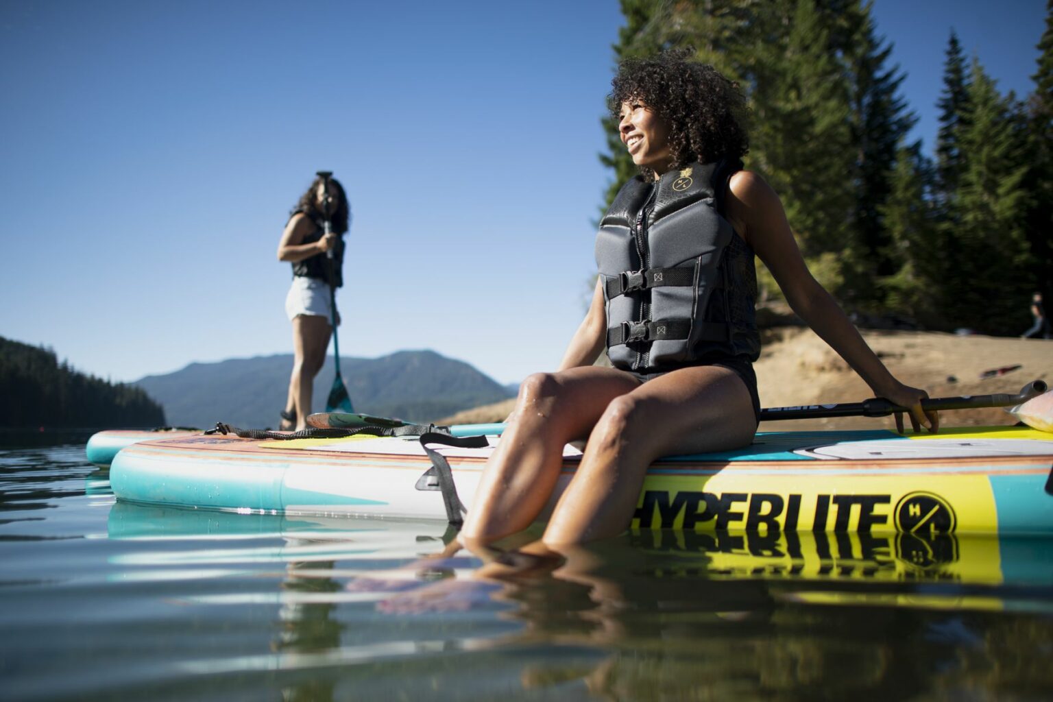 License-free image - Two women enjoy a sunny day on a lake. One is sitting on a paddleboard with her legs in the water, and the other is standing on a standup paddleboard (SUP) in the background. Both wear life vests, with mountains and trees visible in the background.