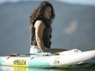 License-free image - A person with curly hair and wearing a life jacket sits on a paddleboard in a calm body of water with distant mountains in the background. The paddleboard has the word &quot;HYPERLITE&quot; on it