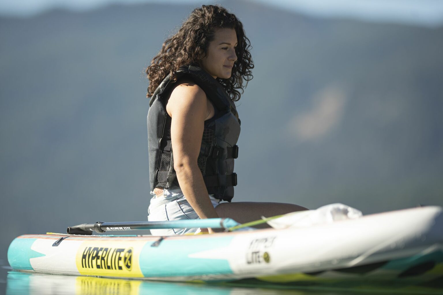 License-free image - A person with curly hair and wearing a life jacket sits on a paddleboard in a calm body of water with distant mountains in the background. The paddleboard has the word &quot;HYPERLITE&quot; on it