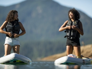 License-free image - Two women are standing on paddleboards in a calm lake, wearing life jackets. They are smiling and looking at each other, with a backdrop of mountain and clear skies. One woman is adjusting her life jacket, and the other is holding onto her paddleboard.