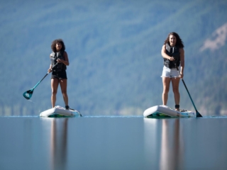 License-free image - Two young women are paddleboarding on a calm lake against a backdrop of distant mountains and trees. They are wearing life vests and appear focused on balancing and paddling on their boards. The water reflects the serene surroundings.