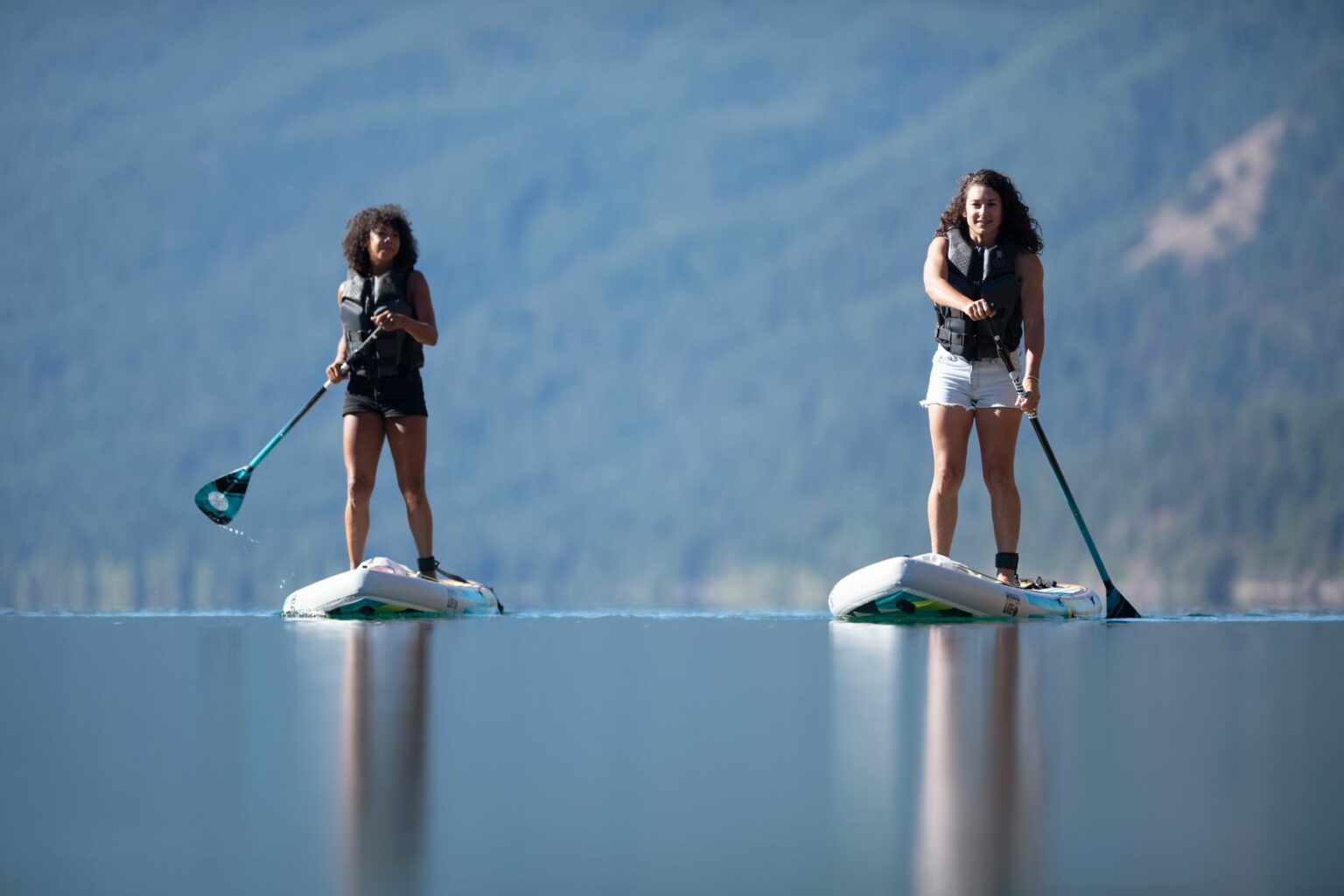 License-free image - Two young women are paddleboarding on a calm lake against a backdrop of distant mountains and trees. They are wearing life vests and appear focused on balancing and paddling on their boards. The water reflects the serene surroundings.