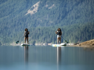 License-free image - Two people are paddleboarding on a calm lake with a mountainous forested background. The weather appears clear and sunny, and the water reflects the surrounding scenery. Both individuals are standing on their boards and paddling.
