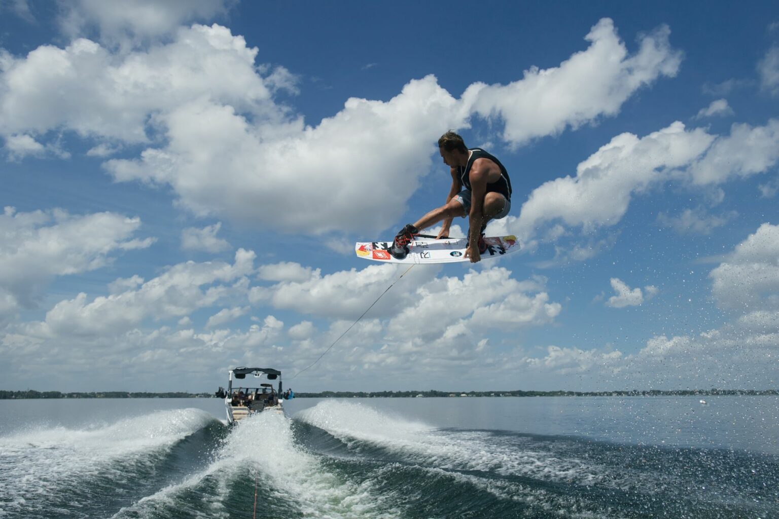 Royalty-free image A person is wakeboarding on a lake, performing a high jump into the air while holding a rope attached to a boat. The boat creates waves in the water, and the sky is filled with scattered clouds on a sunny day.