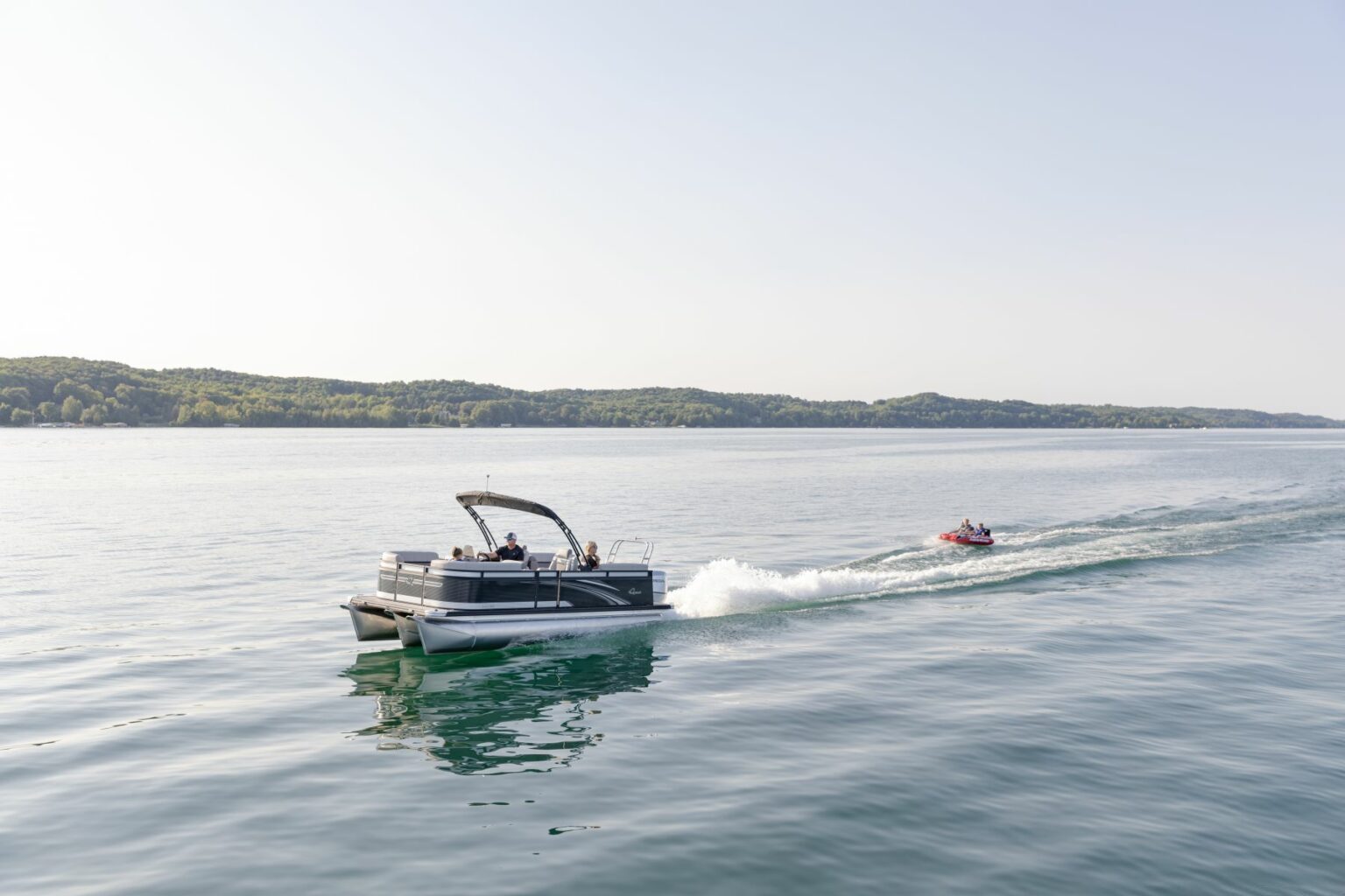 License-free image - A pontoon boat cruising on a calm lake with several people onboard and a person riding a red jet ski nearby. The distant shoreline is covered with trees under a clear sky.