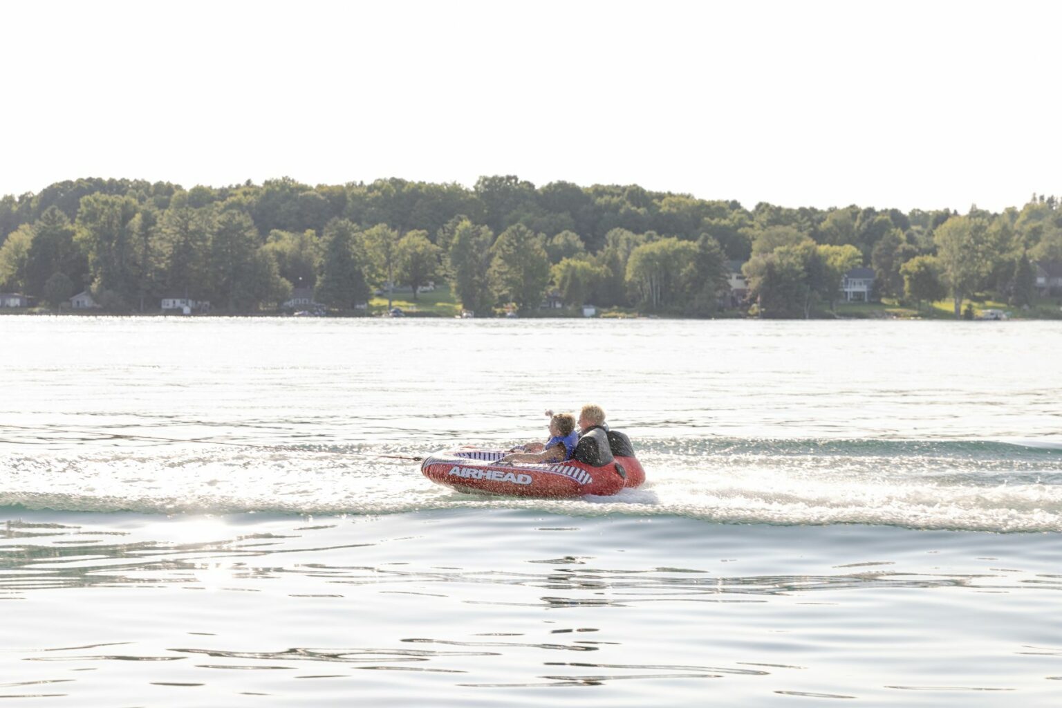 License-free image - Two people are riding an inflatable Airhead tube across a calm lake. The tube leaves a foamy trail on the water as they speed by. Lush green trees and several houses are visible in the background against a bright, slightly overcast sky.