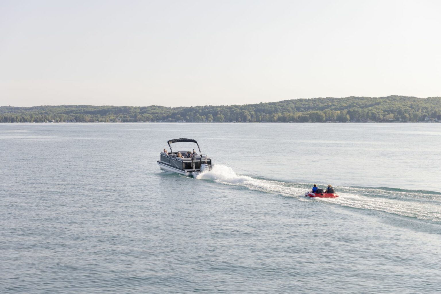 License-free image - A boat speeds across a calm lake towing a person on an inner tube. The water is clear and reflects the light sky. Green forested land is visible in the background under a bright, clear sky.