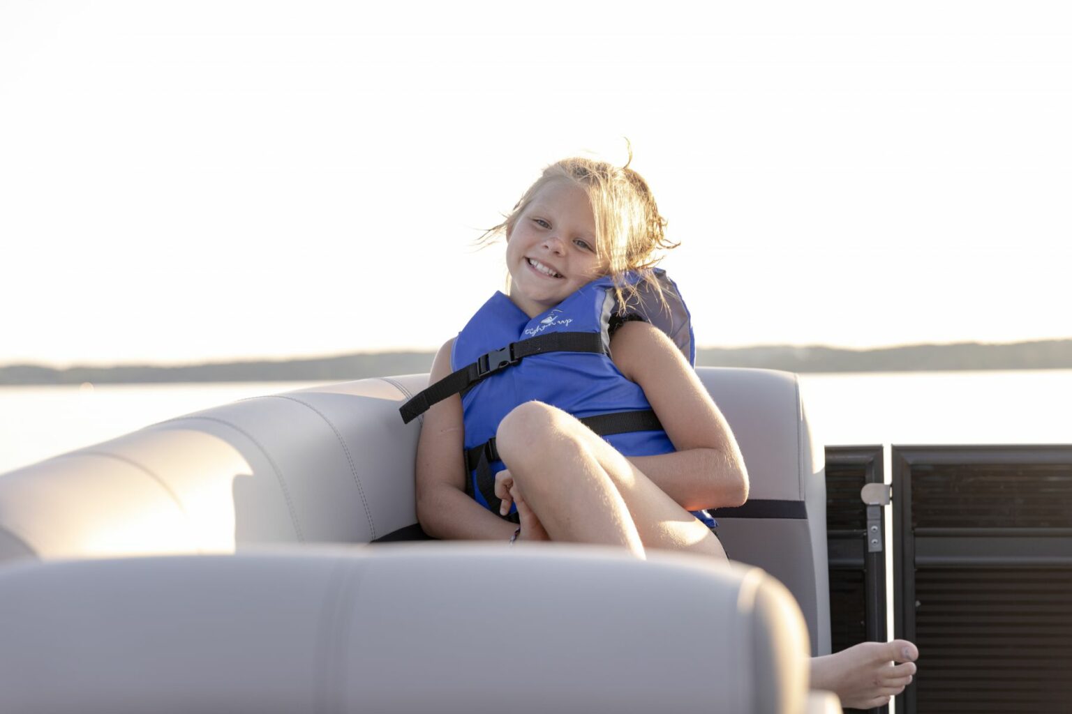 License-free image - A smiling young girl wearing a blue life jacket sits on the seat of a boat. She is relaxed and enjoying the ride on a calm, sunny day. The background shows serene water and a clear sky.