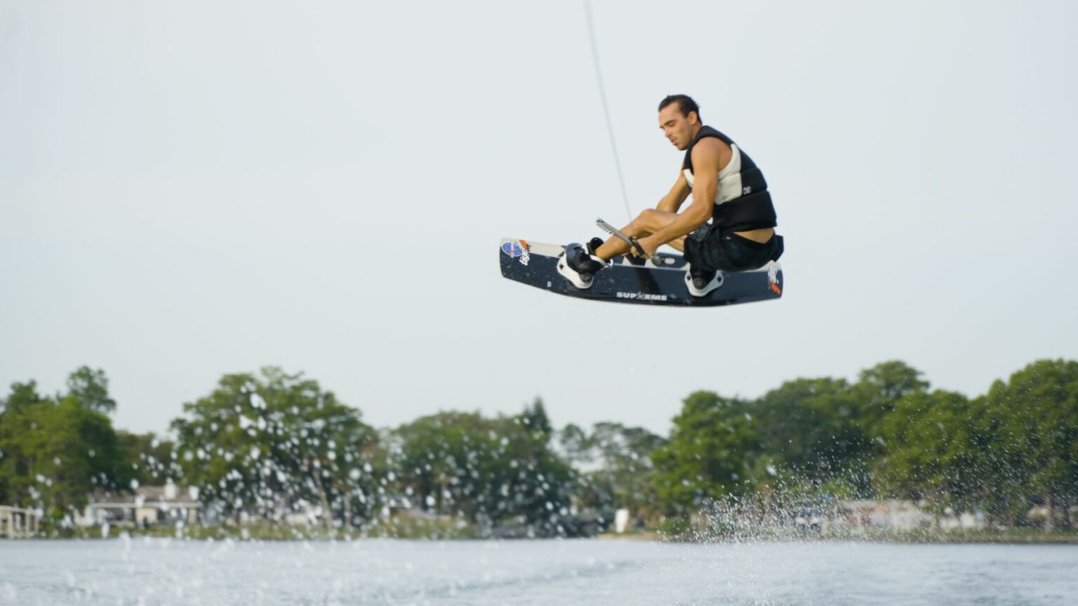 License-free image - A person wearing a black life vest is airborne on a wakeboard above a body of water, with a splash in the foreground. The wakeboarder is holding onto a rope and is high above the water surface, with trees visible in the background.