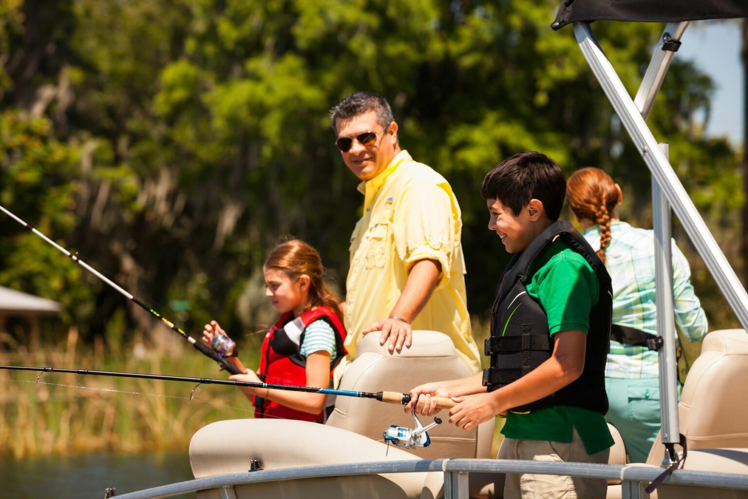 License-free image A family enjoys a day of fishing on a boat. A boy and girl, both wearing life jackets, are casting their fishing rods. A man in sunglasses, likely their father, stands nearby in a yellow shirt. Another person, partially visible, stands at the boat&#039;s helm. Trees are in the background.