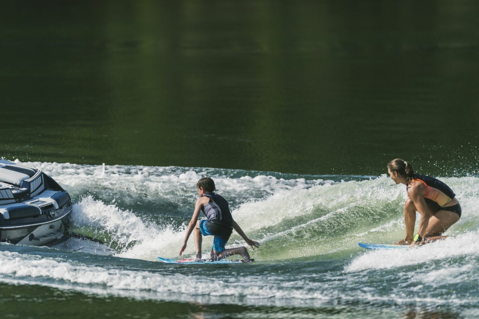 License-free image - Two people are wake surfing on a body of water, being towed by a motorboat. One person, wearing a sleeveless top, shorts, and a life jacket, is surfing close to the boat’s wake, while the other person, in a swimsuit, is surfing behind. The water is calm and green.