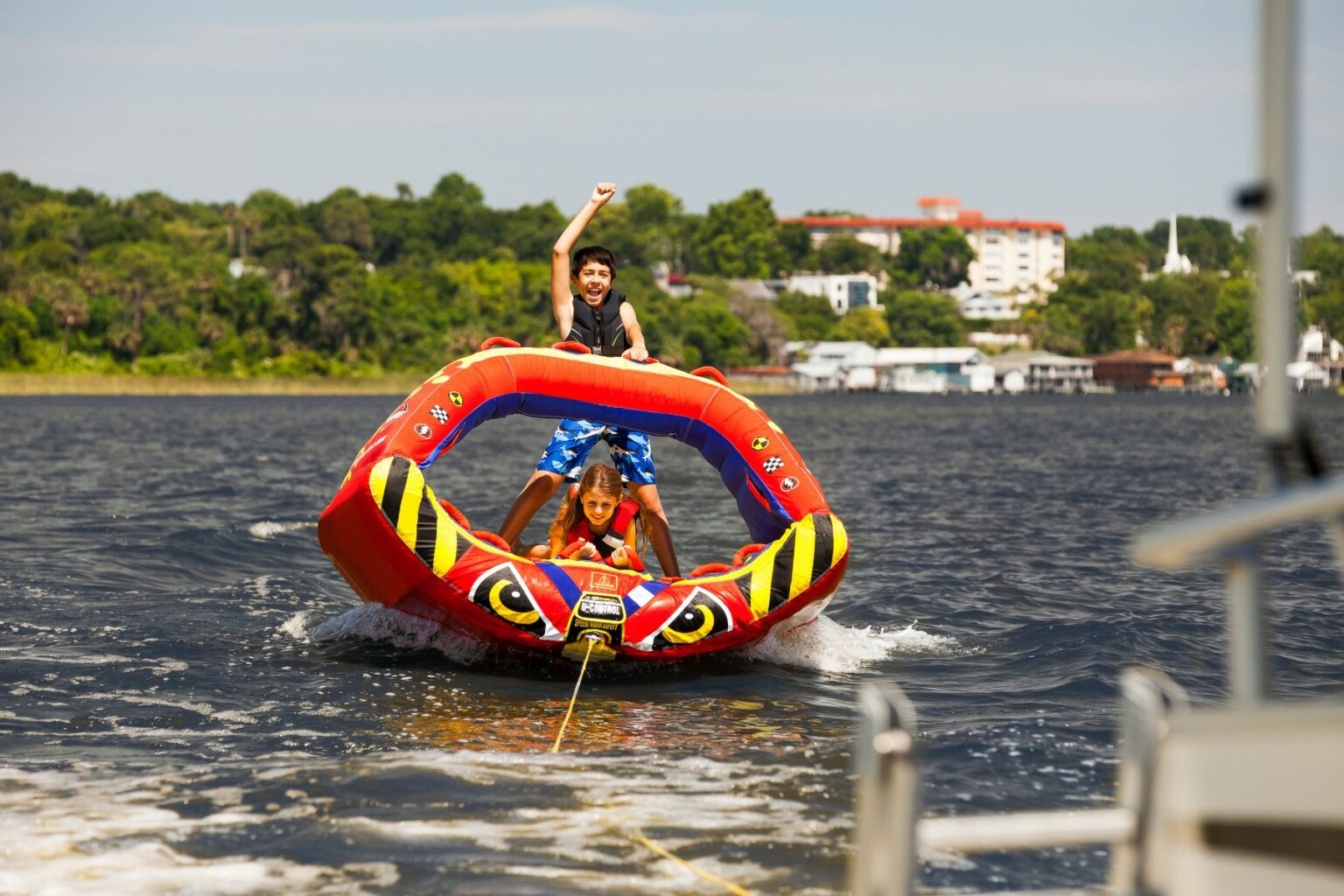 License-free image - Two children ride an inflatable tube being towed by a boat on a sunny day. The child standing raises one arm in excitement, while the other sits holding onto the tube&#039;s handles. Trees and buildings are visible in the background, and wake trails behind the tube.