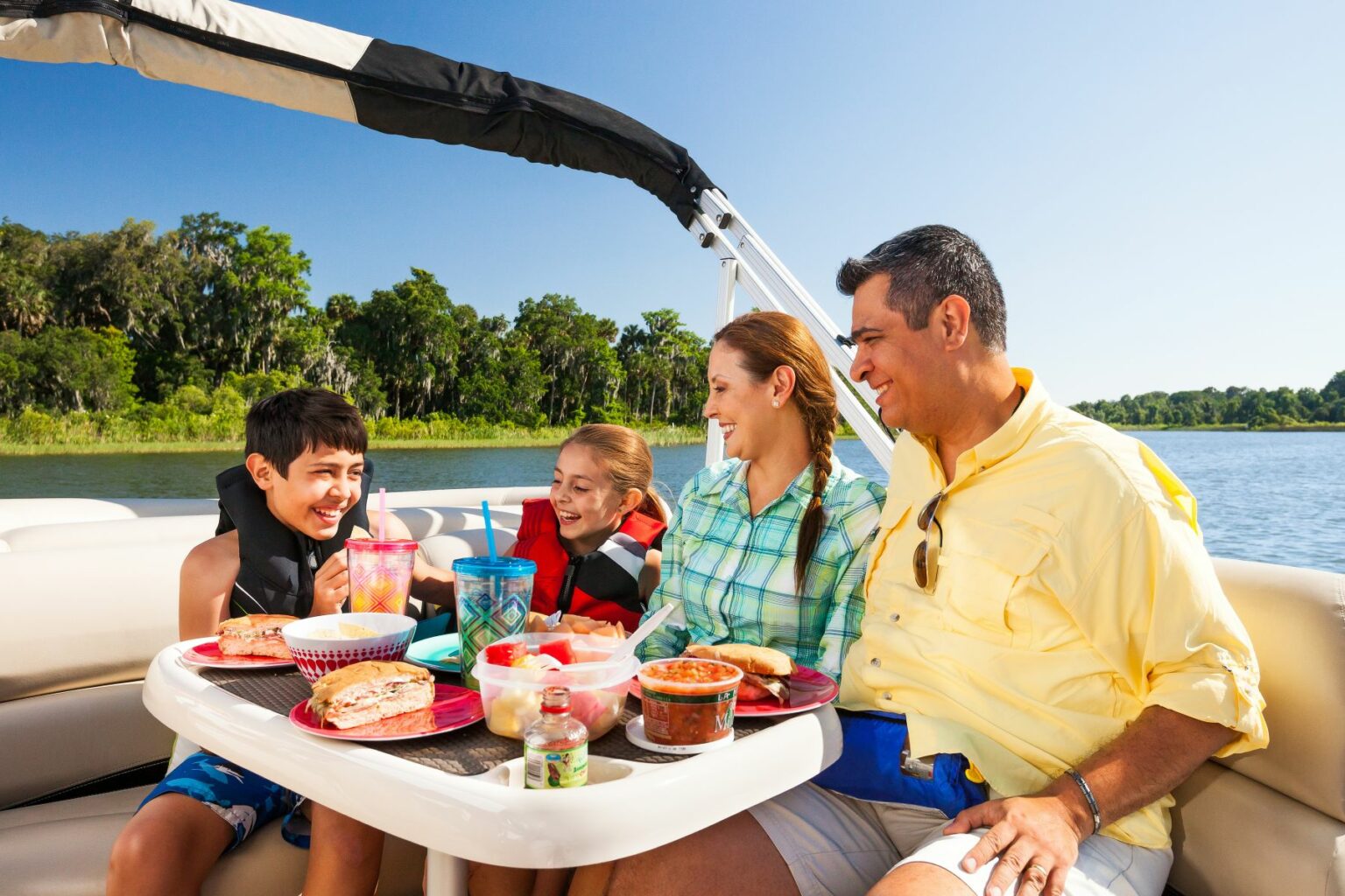 Royalty-free image - A family of four enjoys a sunny day on a boat, sitting at a table with food and drinks. The parents and two children are smiling and wearing casual clothes, with a blue sky and green trees in the background. The children wear life jackets.