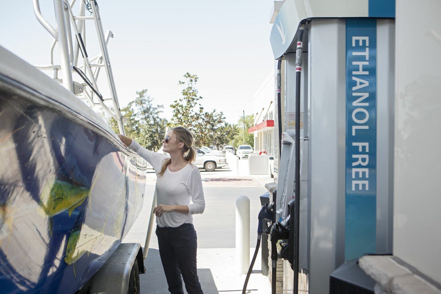 License-free image - A woman wearing sunglasses and a white shirt is pumping ethanol-free fuel into a boat at a gas station. The boat has detailed artwork on its exterior. The fuel pump next to her has a blue sign that reads &quot;ETHANOL FREE&quot;. Trees and a building are in the background. Properly fueling a boat with ethanol free, unleaded gasoline at a land pump.