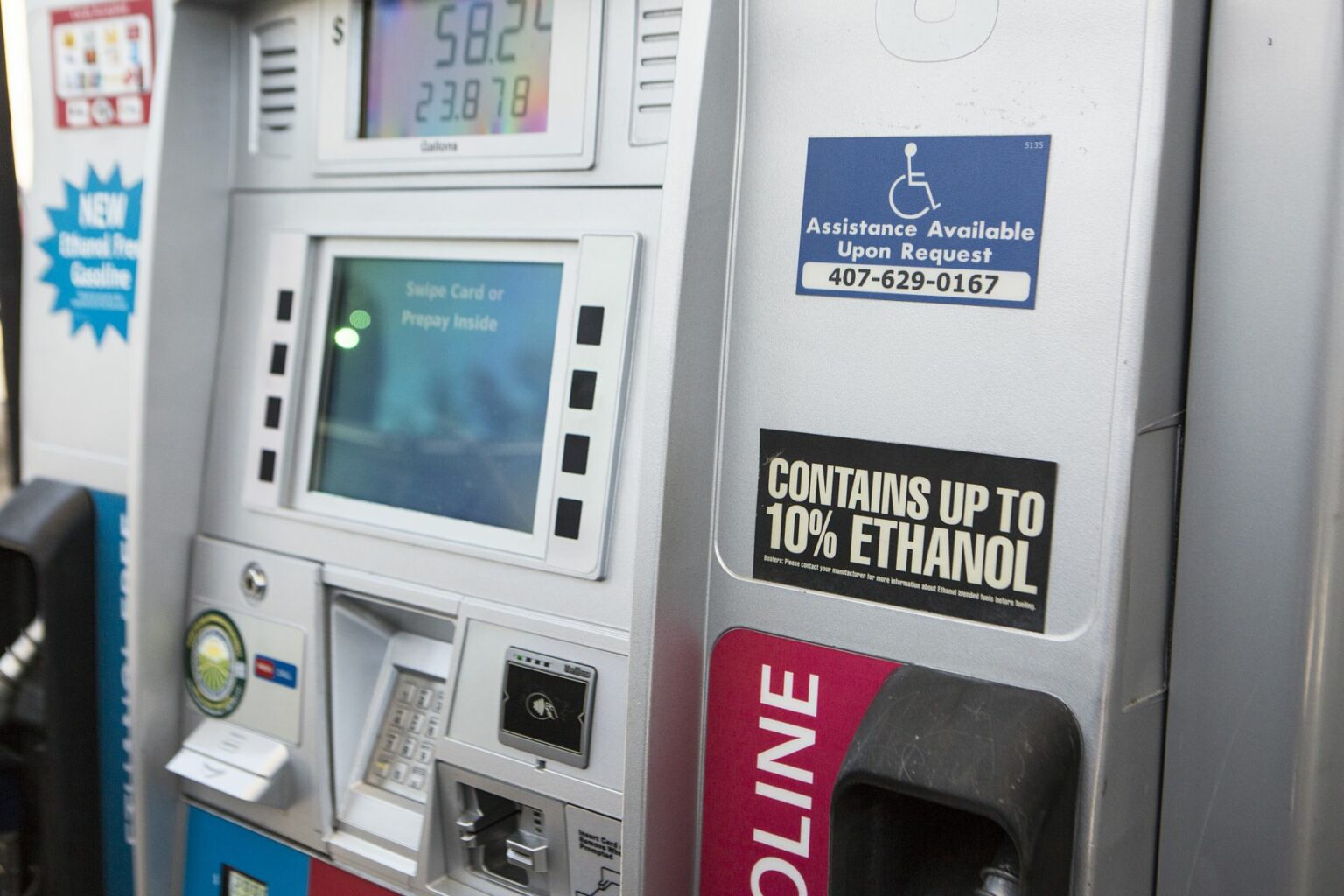 License-free image - Close-up of a gas pump displaying screens, buttons, card swipe area, and a nozzle holder on the left. A sign reads &quot;Contains Up to 10% Ethanol,&quot; and another sign provides a contact number for assistance. A third sign on the left mentions a new payment technology. Gas at a fuel pump with 10% ethanol.