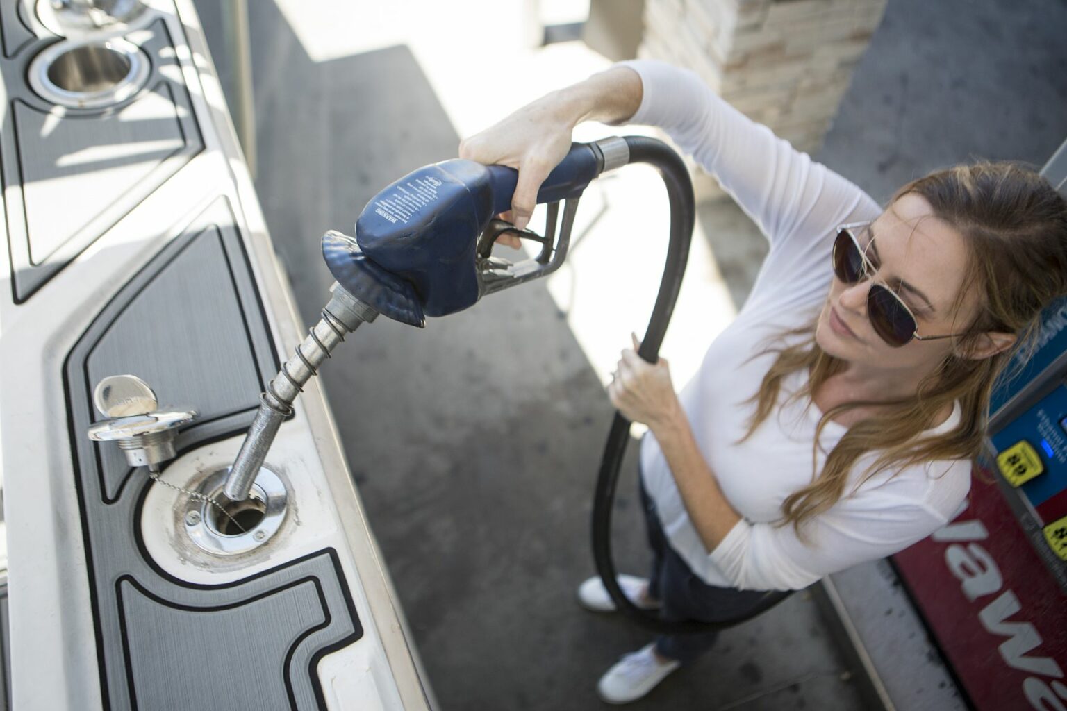 License-free image - A woman dressed in a white long-sleeve shirt and sunglasses is seen from above, holding a fuel nozzle while refueling her boat at a gas station. She appears focused as she inserts the nozzle into the boat&#039;s fuel tank. Inserting the nozzle in the fuel neck for refueling a boat at a land pump.