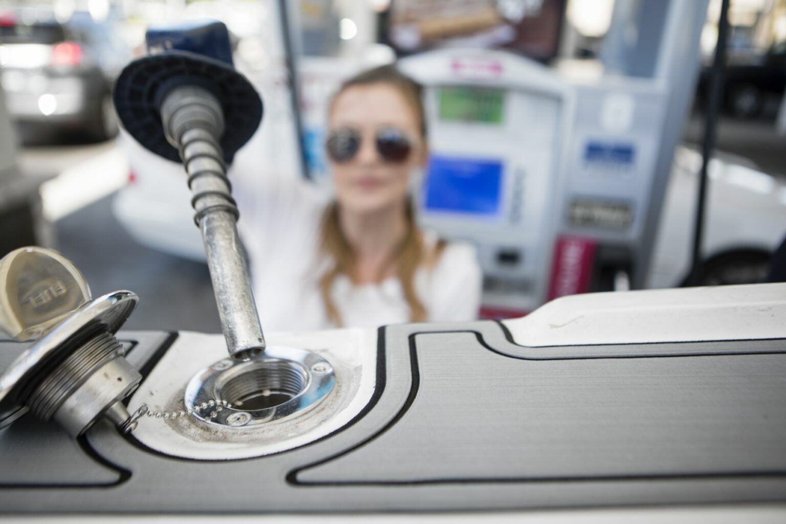 License-free image - A person is seen fueling a vehicle at a gas station. The foreground shows a close-up of the fuel cap and the gas pump nozzle inserted into the fuel tank. The person in the background is blurred, wearing sunglasses. The gas station equipment is also out of focus. Inserting the nozzle in the fuel neck for refueling a boat at a land pump.