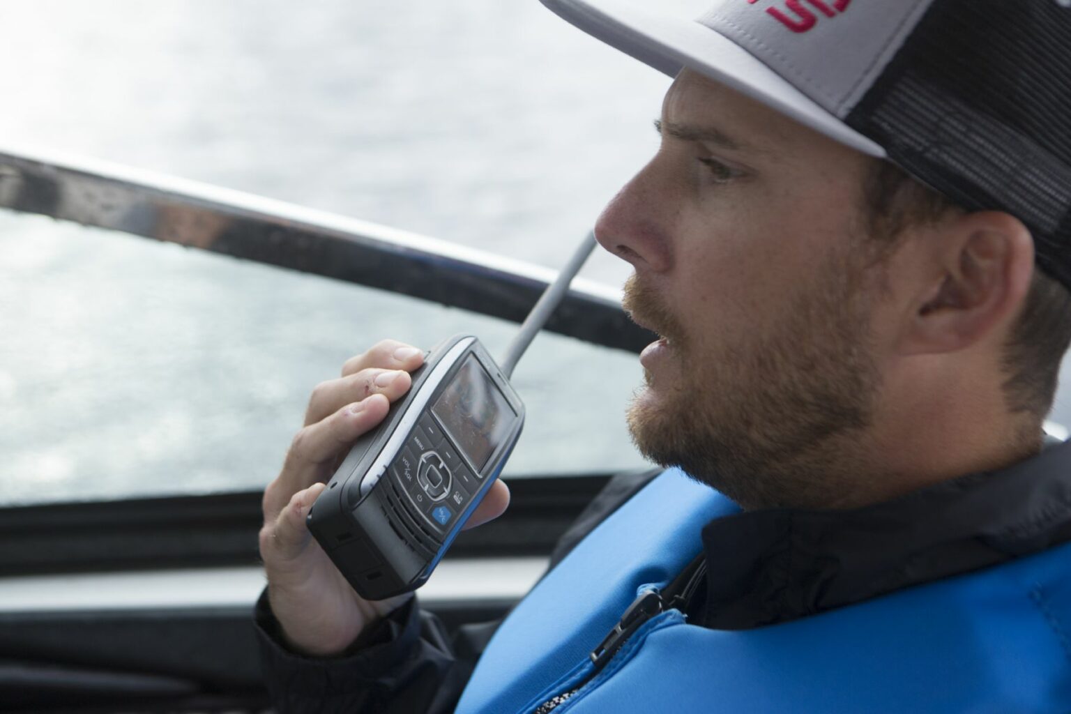 Royalty-free image - A person wearing a blue life vest and a cap is using a handheld VHF-DSC marine band radio set to channel 16 on a boat, speaking while the water shimmers in the background through the window.