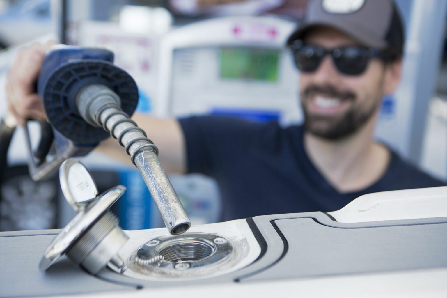 License-free image - A smiling person wearing sunglasses and a cap is holding a fuel nozzle and preparing to refuel a vehicle, with the fuel cap removed and placed beside the open fuel tank. The background shows blurry elements of a gas station. Inserting the nozzle in the fuel neck for refueling a boat at a land pump.