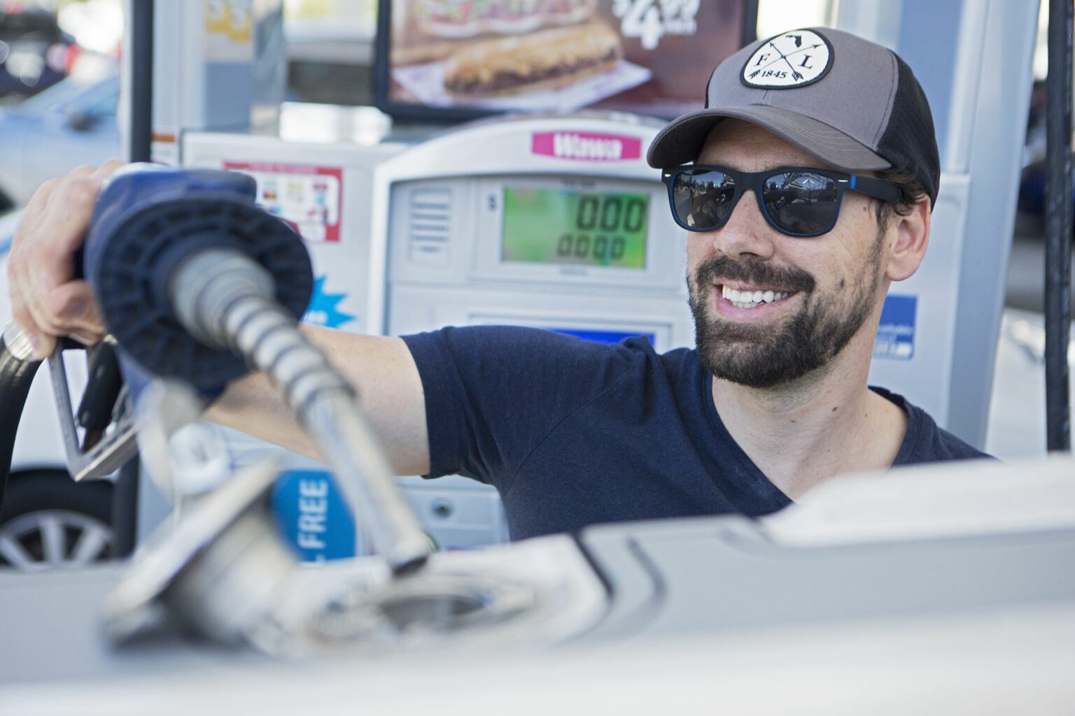 License-free image - A man with a beard, wearing sunglasses and a cap, fills his car with gas at a fuel station. He is smiling and looking towards the gas pump, which displays zeros on the screen. A poster with a food advertisement is visible in the background. Inserting the nozzle in the fuel neck for refueling a boat at a land pump.