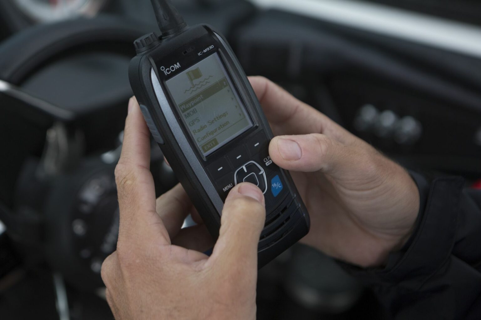 Royalty-free image - Close-up of hands holding and operating a black handheld radio device with a screen displaying navigational information. The background shows part of a steering wheel, indicating a possible marine or vehicular setting. Using a handheld VHF-DSC marine band radio set to channel 16.