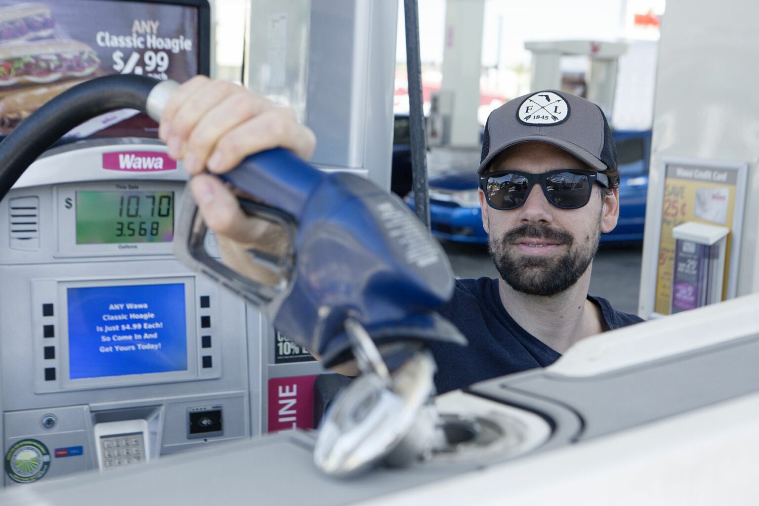 License-free image - A person wearing sunglasses and a hat is seen fueling up a vehicle at a gas station. They are holding a blue fuel nozzle at the opening of the vehicle&#039;s gas tank. Price and product advertisements are visible in the background. Refueling a boat at a land pump.