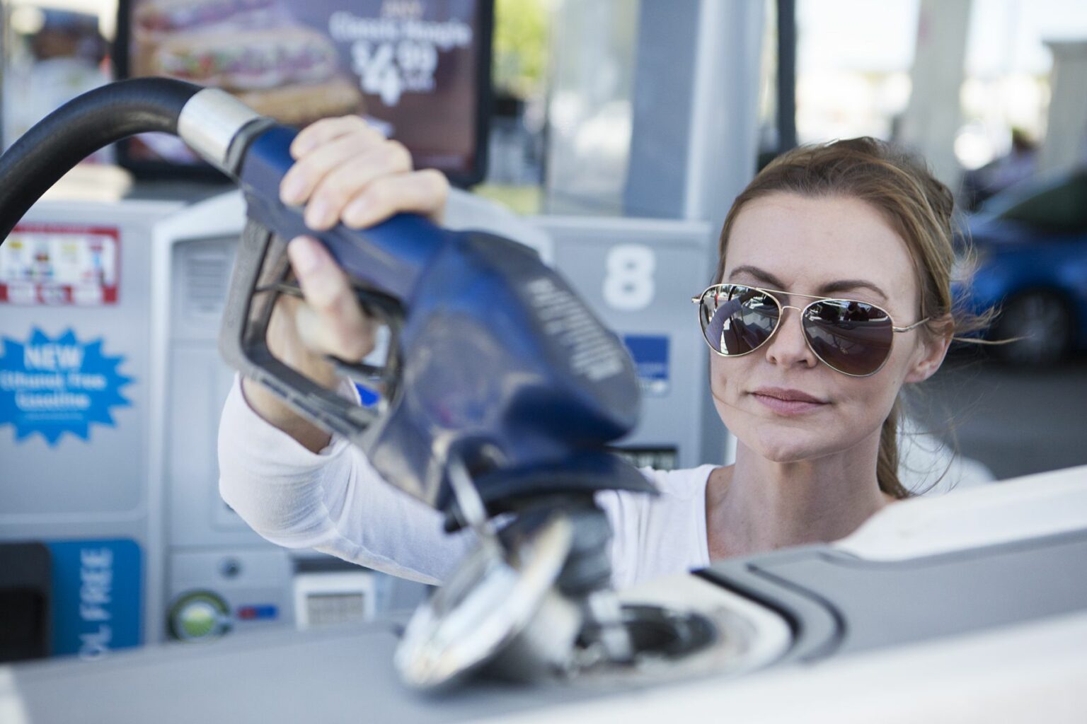 License-free image - A woman wearing sunglasses and a white shirt refuels her car at a gas station. She is lifting the fuel nozzle to insert it into the car&#039;s fuel tank. Gas station signage and a partially visible blue car are in the background. Refueling a boat at a land pump.