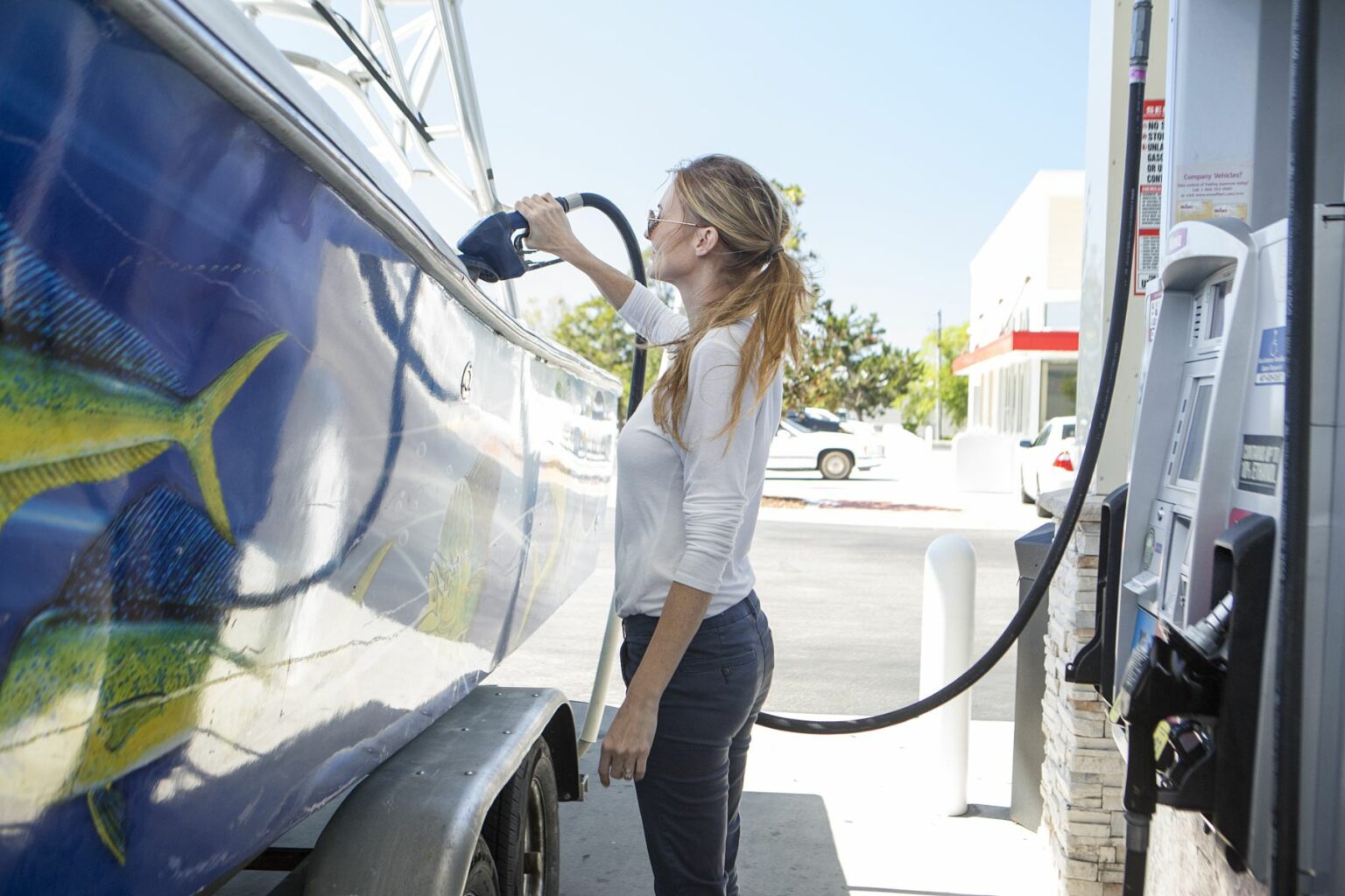 License-free image - A woman is refueling a boat with colorful fish art on its side at a gas station. She is holding a fuel nozzle and wearing a casual white long-sleeve shirt and sunglasses. The background shows part of the gas station and a car, with a clear blue sky above. Refueling a boat at a land pump.