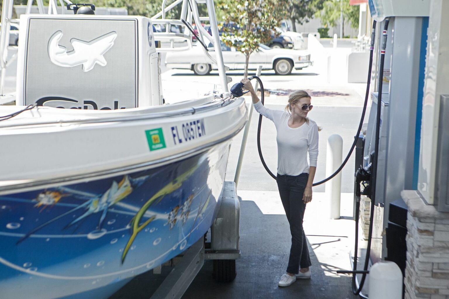 License-free image - A woman in sunglasses and a white shirt is fueling a boat at a gas station. The boat features colorful aquatic artwork on its side, and the pump hose is connected to the boat. Trees and parked vehicles are visible in the background. Refueling a boat at a land pump.