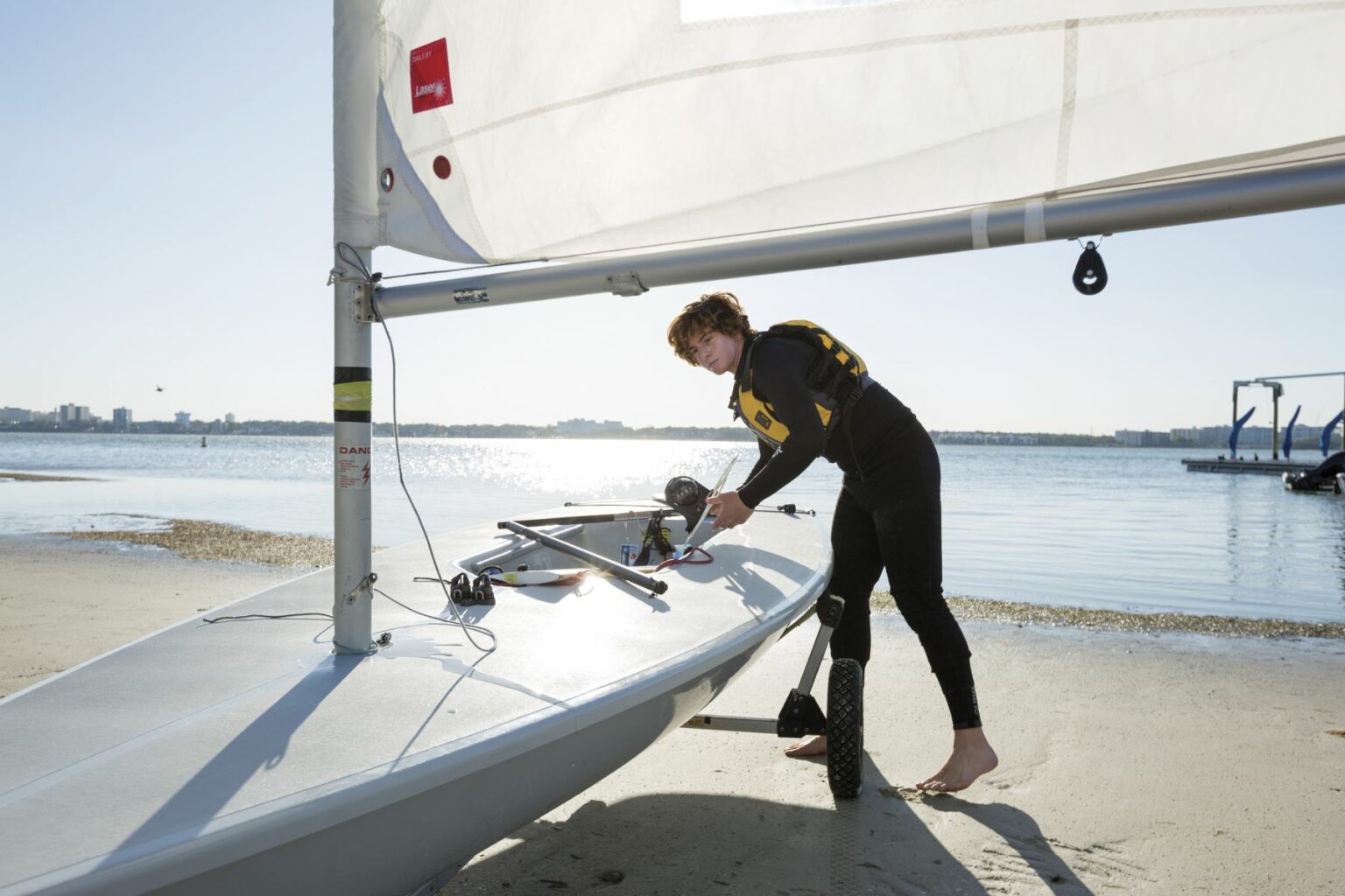 License-free image - A person wearing a yellow life vest and black wetsuit prepares their sailboat on a sandy beach. The person is adjusting the boat&#039;s rigging near a calm body of water, with a cityscape visible in the background. The sky is clear and it appears to be a sunny day. Rigging the sail while wearing a properly fitting life vest in the Sand Key inlet in Clearwater Beach.