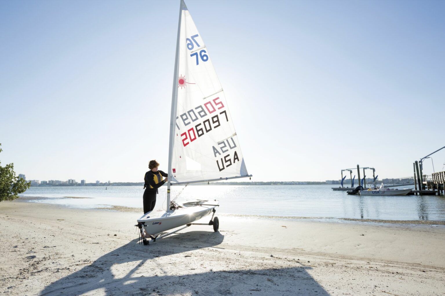 License-free image A person stands next to a small sailboat labeled &quot;206097 USA&quot; on a sandy beach. The person, dressed in a black wetsuit, faces the water with trees and a docked boat visible in the background under a clear blue sky. Preparing to sail while wearing a properly fitting life vest in the Sand Key inlet in Clearwater Beach.