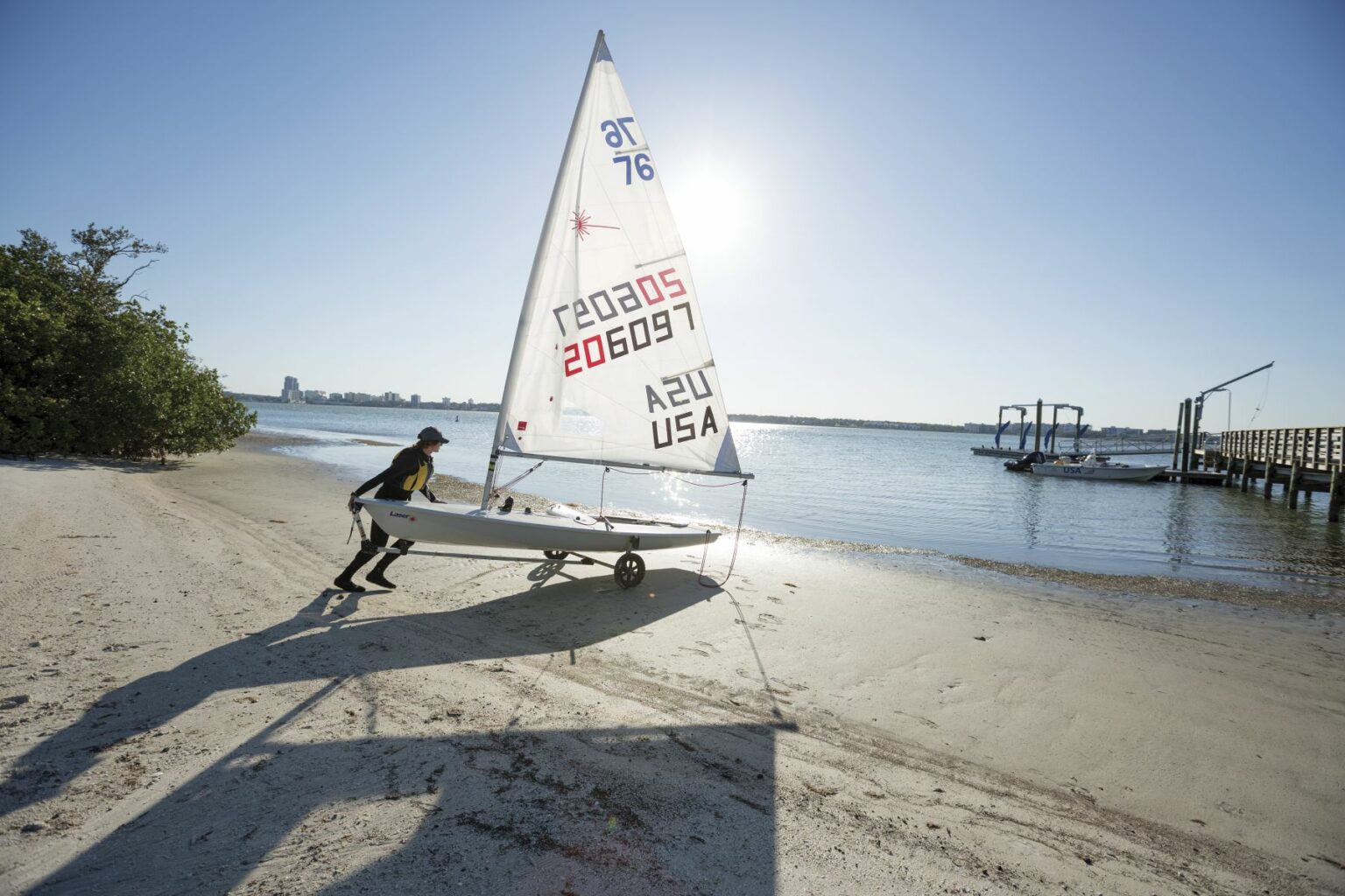 License-free image - A person is pushing a small sailboat on a trailer towards the water on a sunny day. The sailboat has a white sail with identifiable numbers and &quot;USA&quot; written on it. Nearby, there is a wooden dock, trees, and buildings in the distant background. Preparing to sail while wearing a properly fitting life vest in the Sand Key inlet in Clearwater Beach.