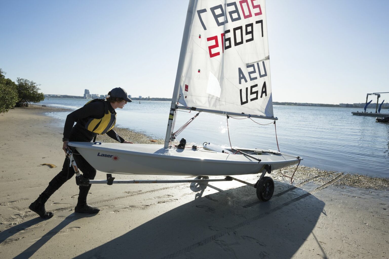 License-free image - A person wearing a life jacket and cap is pulling a small sailboat on a wheeled dolly from the shore into the water. The sailboat has &quot;LASER&quot; and &quot;USA&quot; written on the sail along with the number &quot;210597.&quot; The background features water, greenery, and distant buildings. Sailing while wearing a properly fitting life vest in the Sand Key inlet in Clearwater Beach.