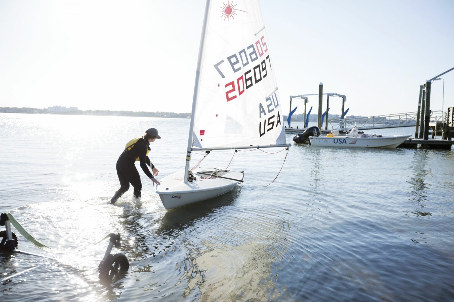 License-free image A person wearing a wetsuit prepares to set sail on a small sailboat marked with &quot;206907&quot; and &quot;USA&quot; at a dock. The boat is partially in the water, and a metal launching trolley is visible in the foreground. Bright sunlight reflects off the water&#039;s surface. Sailing while wearing a properly fitting life vest in the Sand Key inlet in Clearwater Beach.